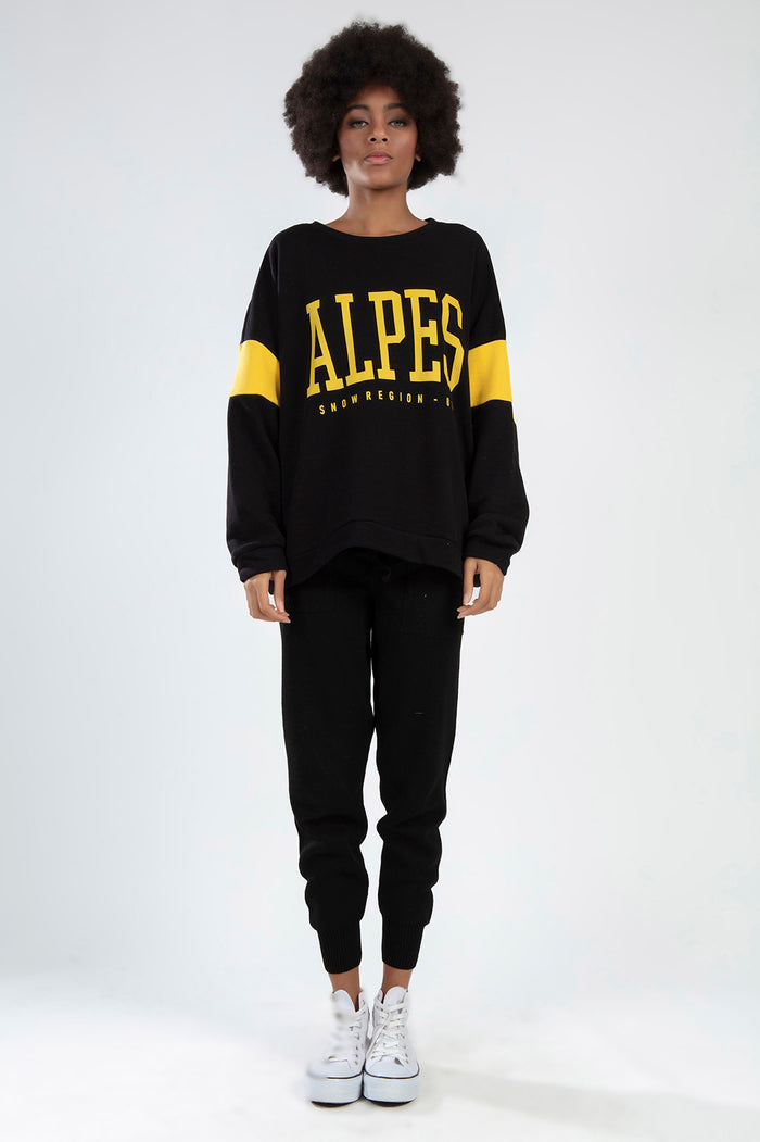 Model in black ALPES streetwear crop top with yellow graphics and stripe sleeves, paired with matching ribbed joggers.