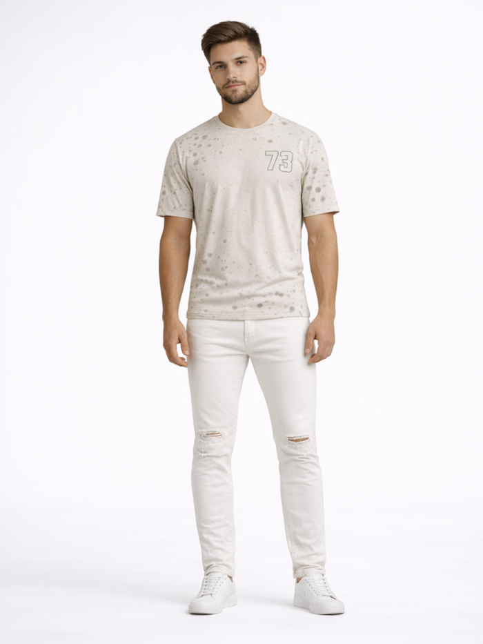A man stands confidently in light-colored ripped jeans and a speckled t-shirt, paired with white sneakers.