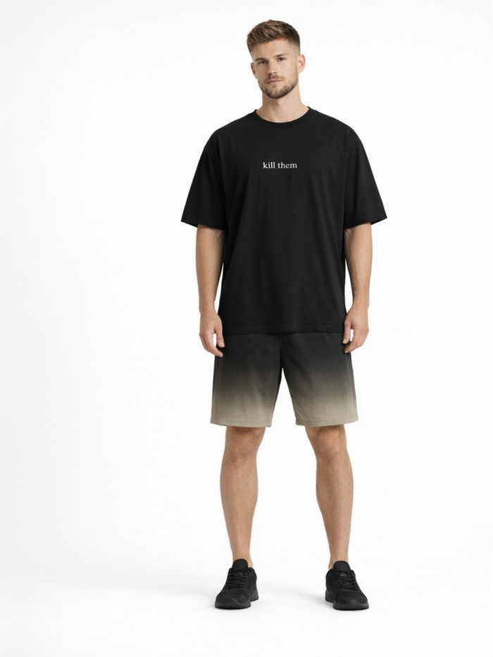 A man stands wearing a black oversized t-shirt with white text, paired with gradient shorts and black sneakers.