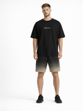A man stands wearing a black oversized t-shirt with white text, paired with gradient shorts and black sneakers.