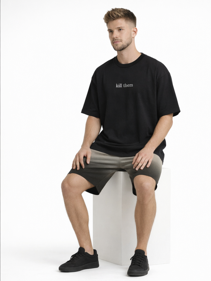 A man sits on a white pedestal wearing a black oversized t-shirt and dark shorts, paired with black sneakers.