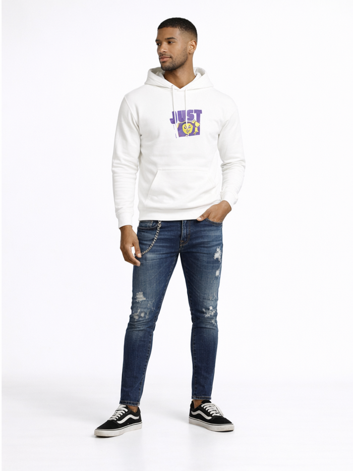 A man stands wearing a white hoodie with a graphic design, paired with distressed blue jeans and black sneakers.
