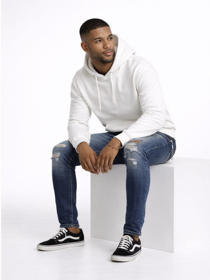 A man sits on a white pedestal wearing a light hoodie, distressed jeans, and black sneakers, looking thoughtfully to...