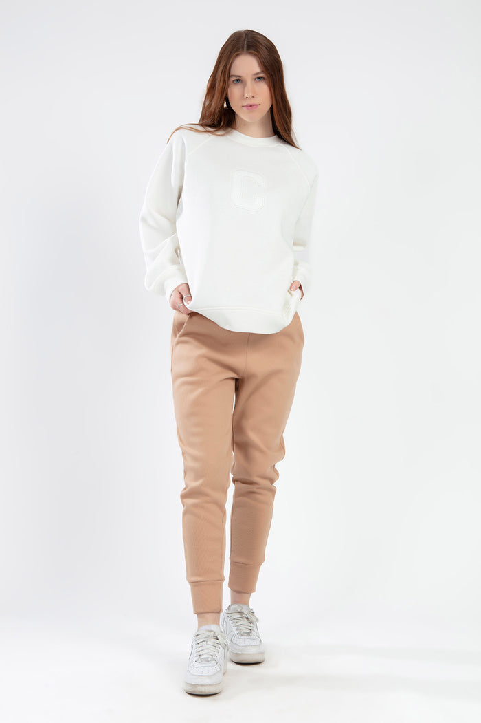 Model in cream crewneck sweatshirt with 'C' logo and tan jogger sweatpants. Trendy Gen Z streetwear loungewear set.