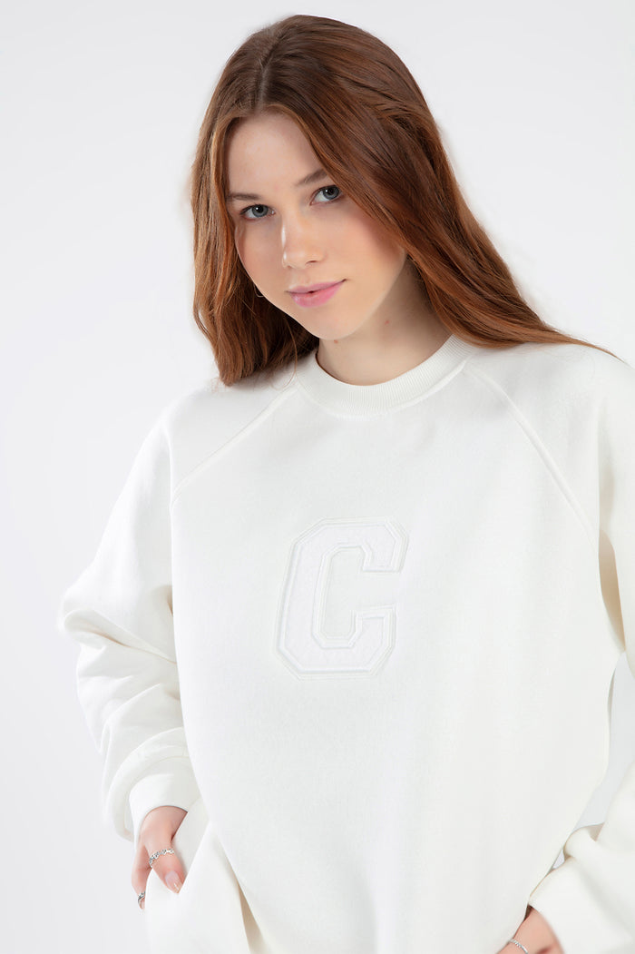 Close-up of a woman wearing a cream crewneck sweatshirt with a tonal embroidered C logo on the chest.