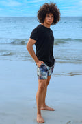 Man on beach wearing Carter patterned swim trunks and black t-shirt, showing trendy summer swimwear style.