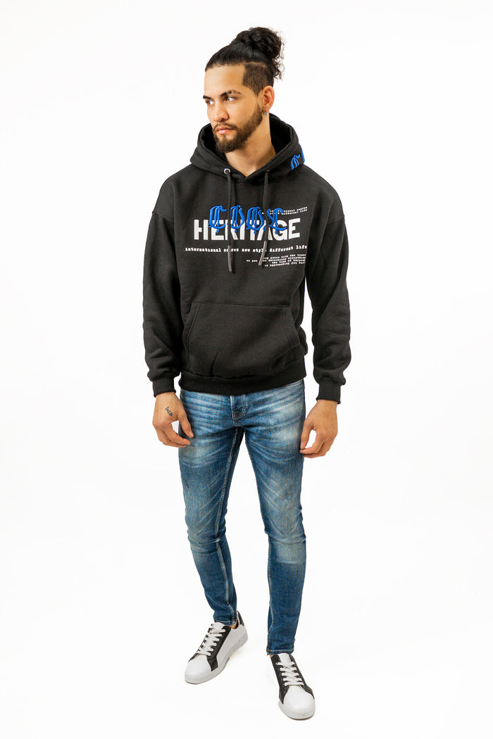 Men's oversized black streetwear hoodie with 'Heritage' blue embroidery, paired with blue jeans and white sneakers.