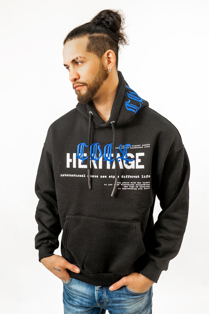 Model wearing a black oversized streetwear hoodie with blue embroidered 'COOL' and white 'HERITAGE' graphic.