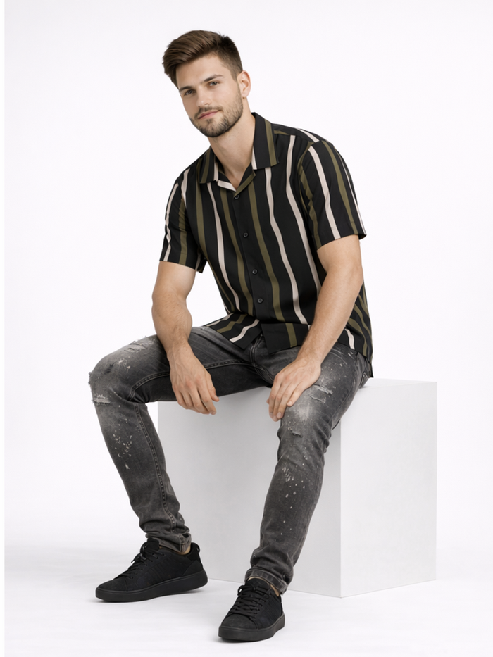 A man sits on a white cube, wearing a short-sleeve striped shirt with dark and light tones, paired with distressed je...