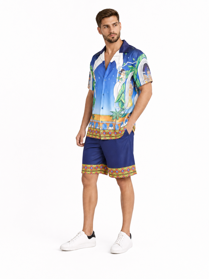 A man stands wearing a colorful short-sleeve shirt with a tropical design and matching blue shorts.