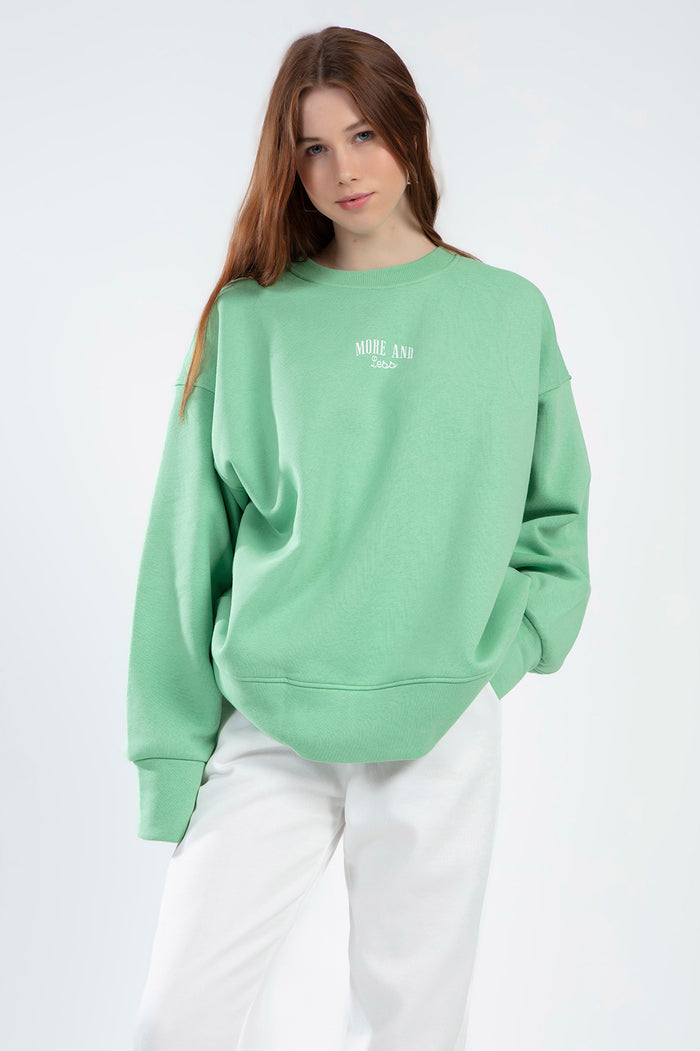 Model wearing the Eve Top, an oversized light green crewneck sweatshirt with 'More and Less' embroidered on the chest.