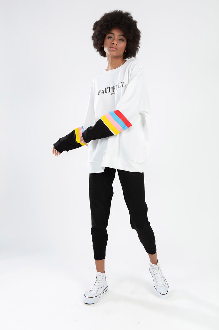 Model wearing FAITHFUL white oversized sweatshirt with rainbow striped sleeves and black joggers.