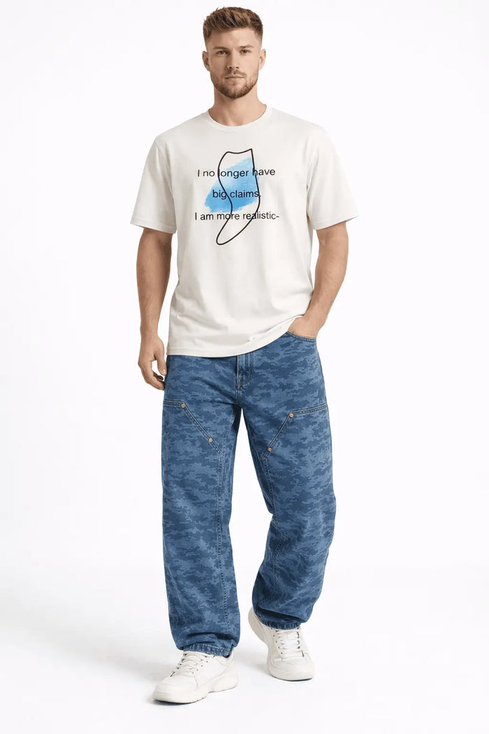 A man stands casually wearing a graphic t-shirt and loose-fitting camo pants, paired with white sneakers.