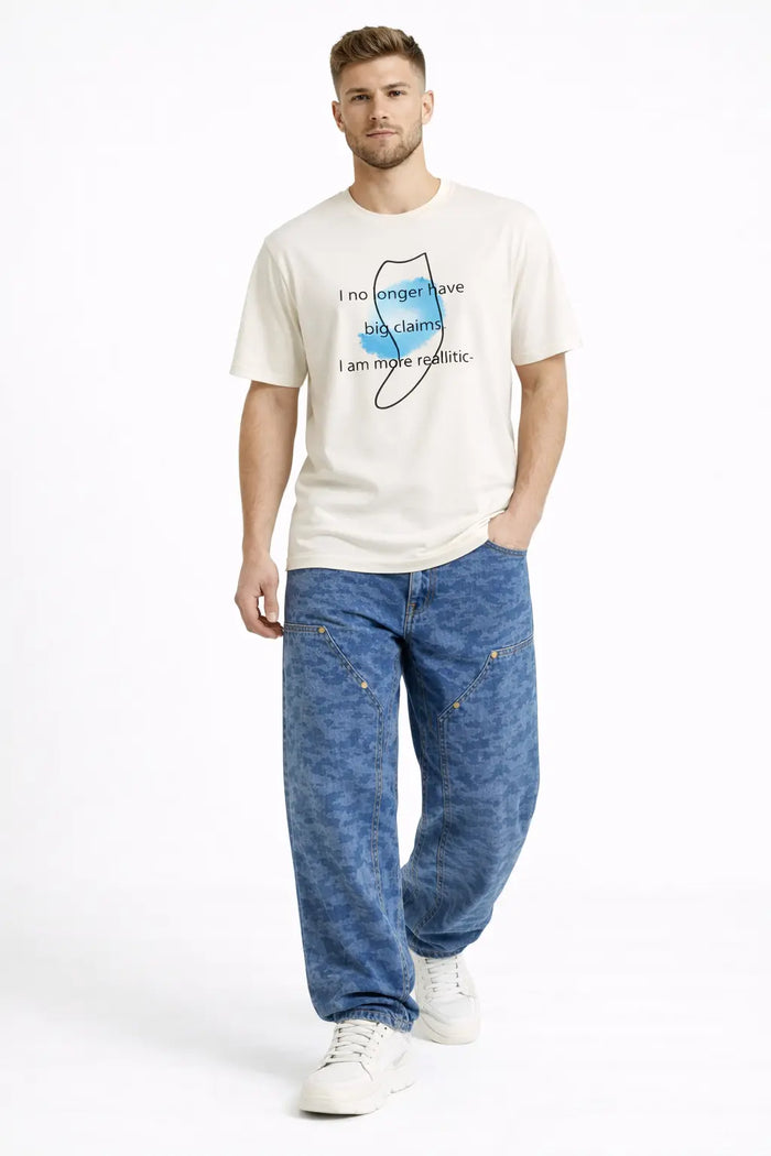 A man wearing a cream graphic t-shirt with blue text and relaxed blue patterned jeans, paired with white sneakers.