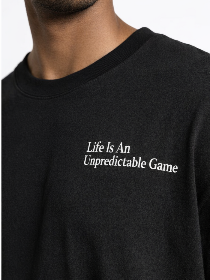 A close-up of a black T-shirt featuring white text that reads "Life Is An Unpredictable Game" on the chest.