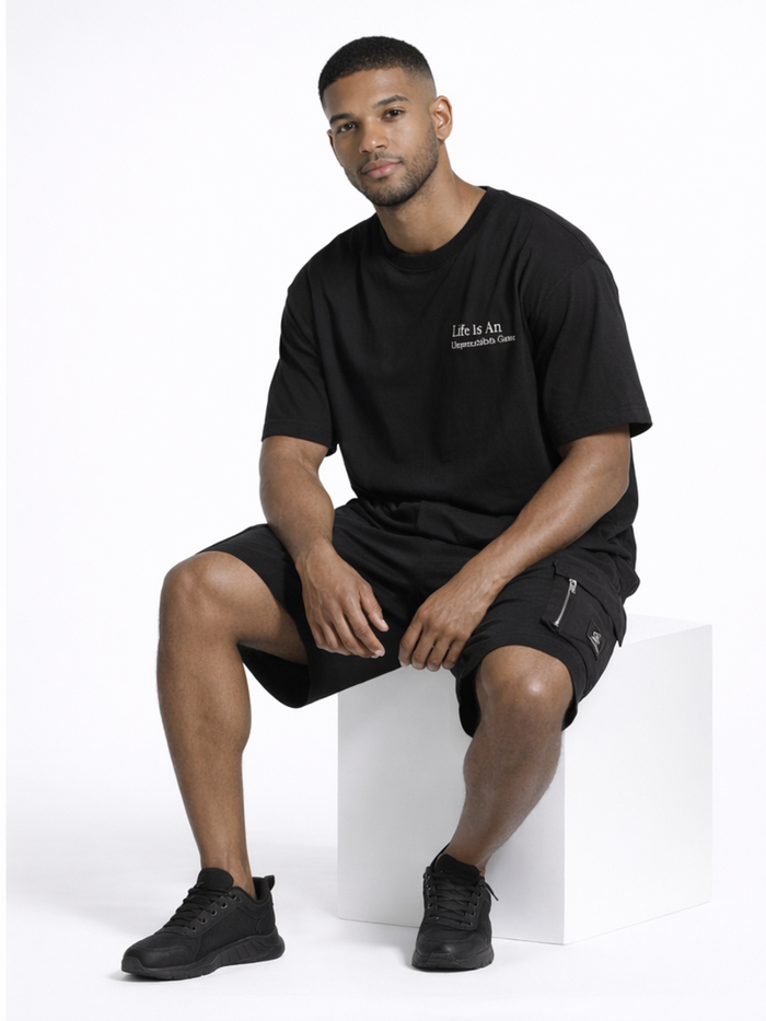 A man sits on a white cube wearing a black oversized t-shirt and black shorts, paired with black sneakers.