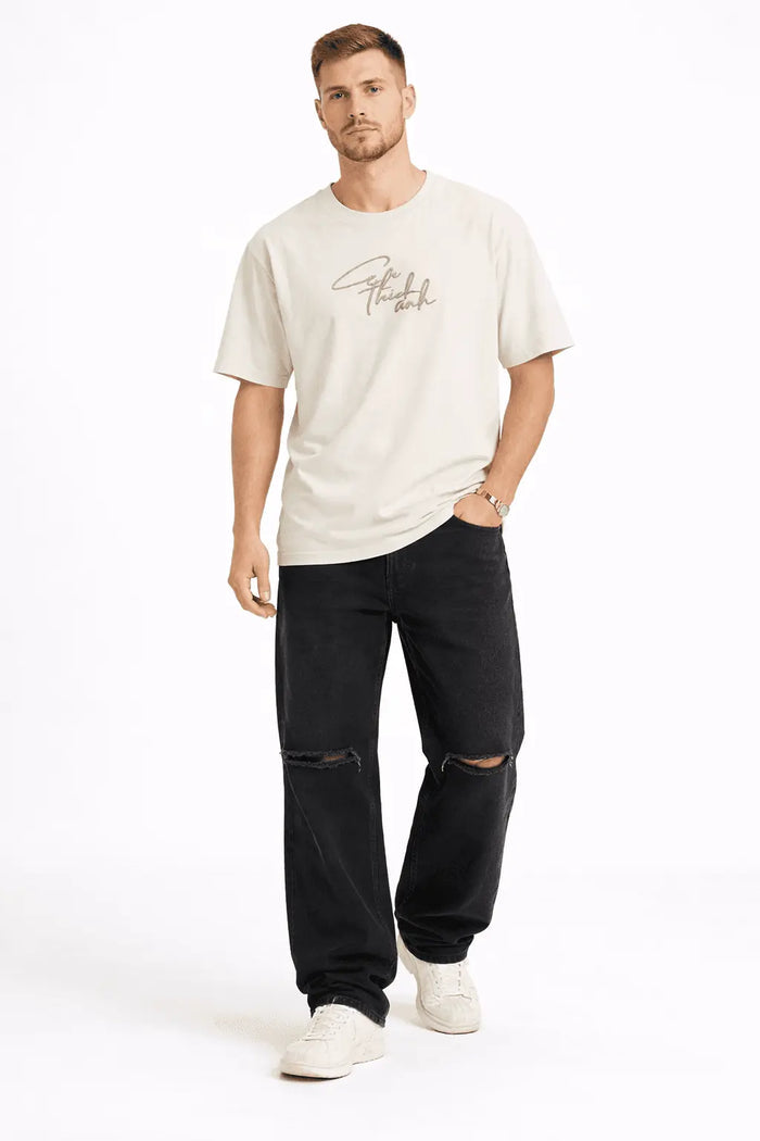 Man wearing Black Baggy Fit Denim streetwear jeans with knee distressing, a cream graphic tee, and white sneakers.