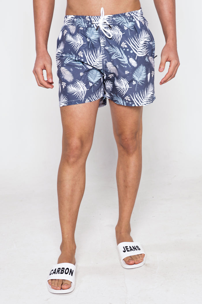 Men's blue Hank swim trunks with white tropical palm leaf pattern, drawstring waist, and Carbon Jeans white slides.