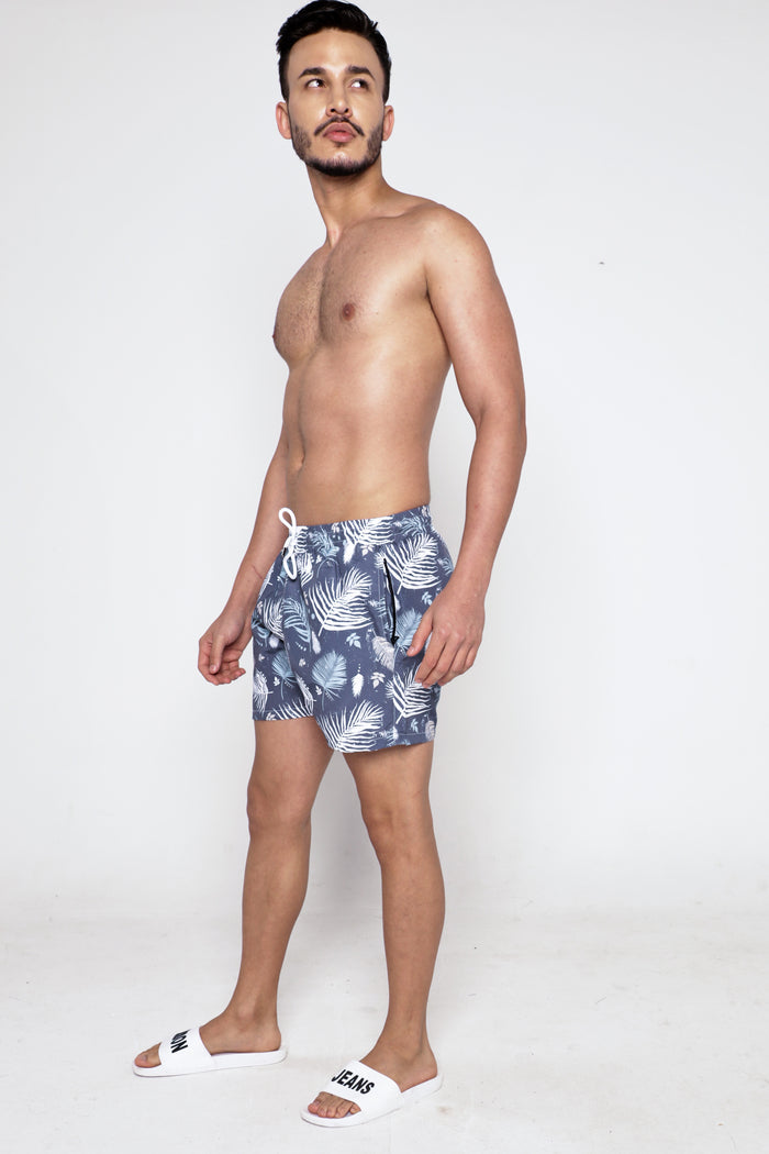 Model wearing blue Hank Swimwear patterned swim trunks with white palm leaf prints and drawstring elastic waistband.