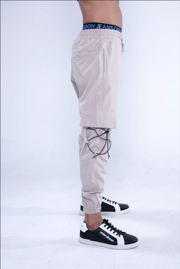 Side view of tan Jesus Cargo Pants with adjustable black bungee cords and elastic cuffs, paired with black sneakers.