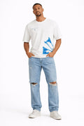 A man stands confidently wearing a white graphic t-shirt with blue designs and light blue distressed jeans.