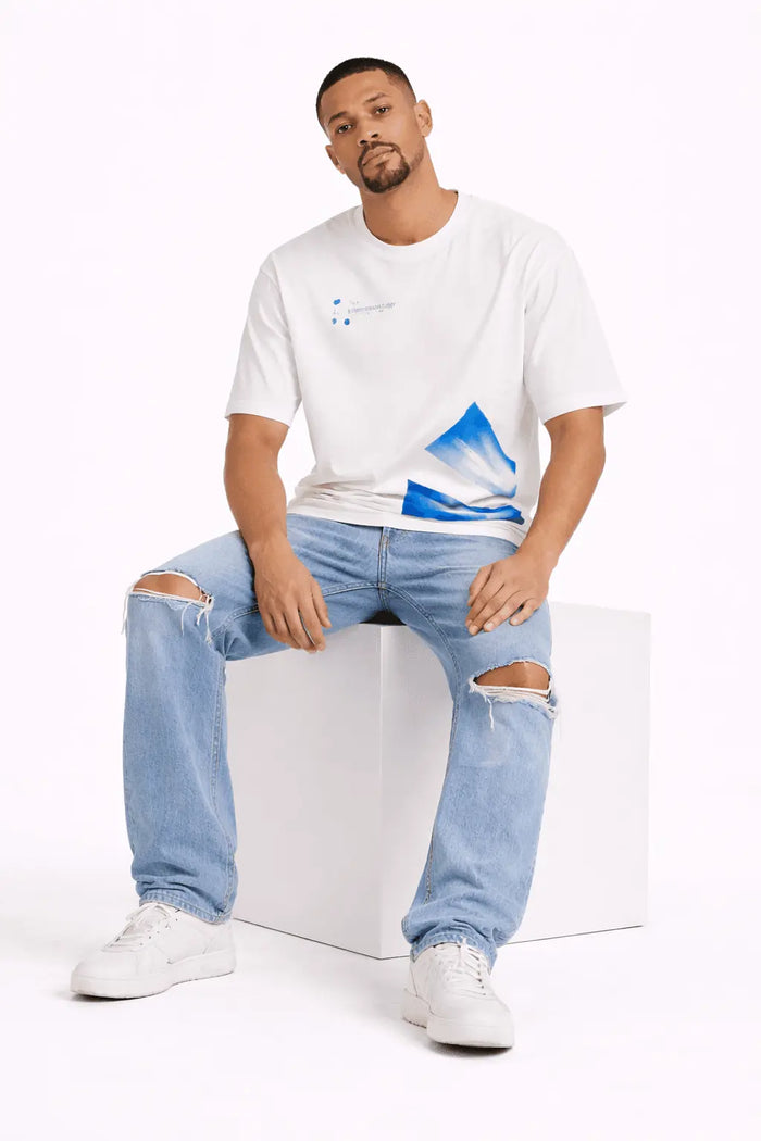 A man sits on a white cube wearing a white t-shirt with blue graphic accents and distressed light blue jeans.