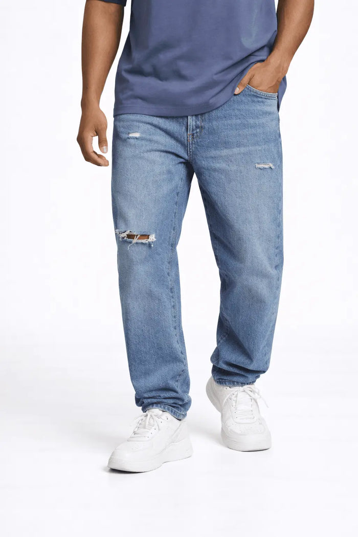 A person wearing light blue distressed jeans with a relaxed fit and white sneakers, standing casually.