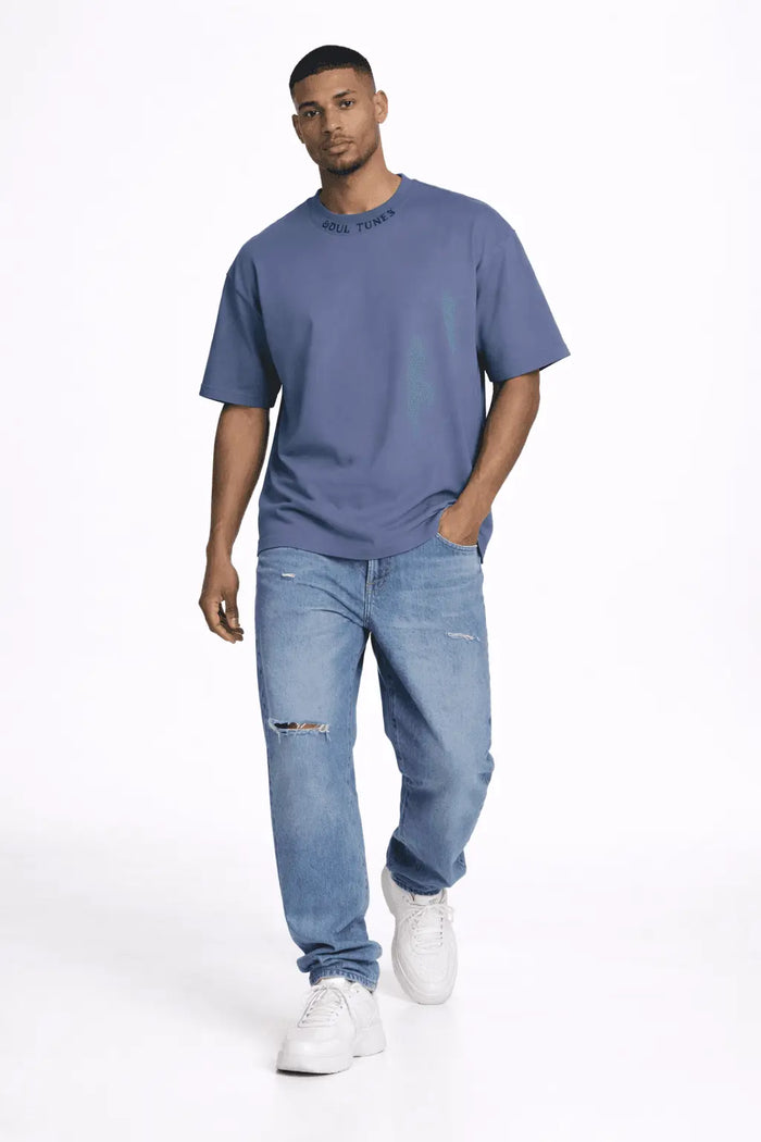 A man walks confidently in a loose-fitting blue shirt and distressed jeans, paired with white sneakers.
