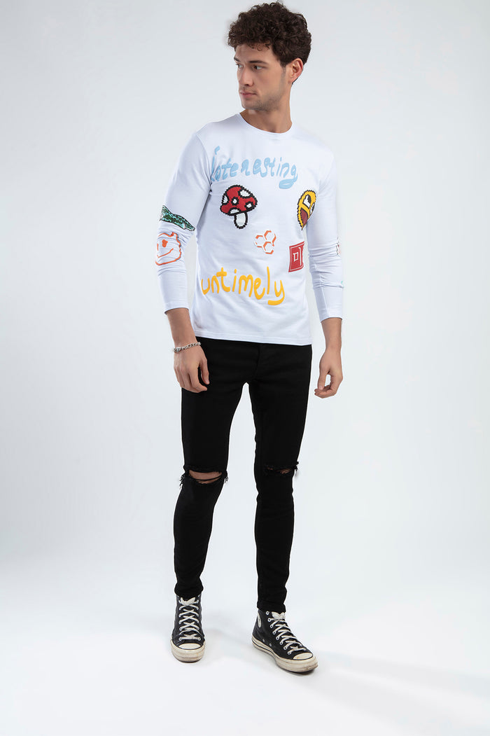 A model wearing a long-sleeve white shirt with colorful graphics and black ripped jeans, paired with sneakers.