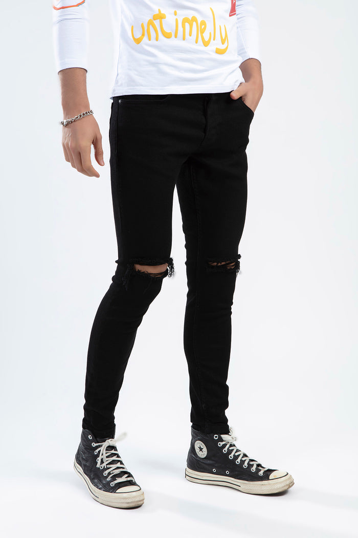 A person wearing black ripped skinny jeans and black high-top sneakers, with a white long-sleeve shirt.