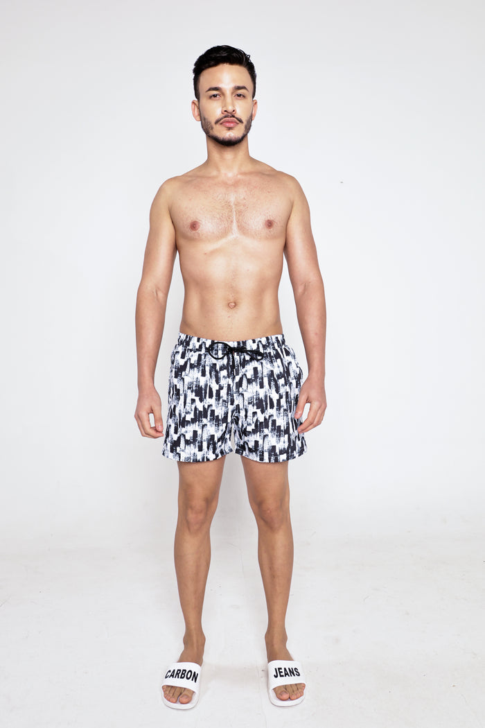 A man wearing Leonardo black and white patterned swim trunks with a drawstring waist and Carbon Jeans slides.