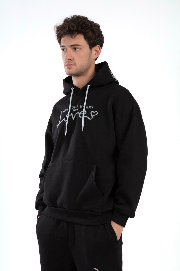 Man in a black oversized Love Hoodie featuring white 'Fuel Your Heart With Love' script embroidery and street style fit.