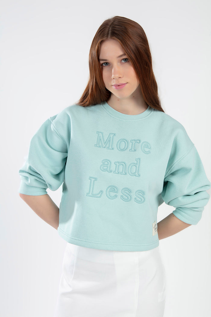 Model wearing a teal knit More and Less cropped sweatshirt with embossed text and puff sleeves against a white background.