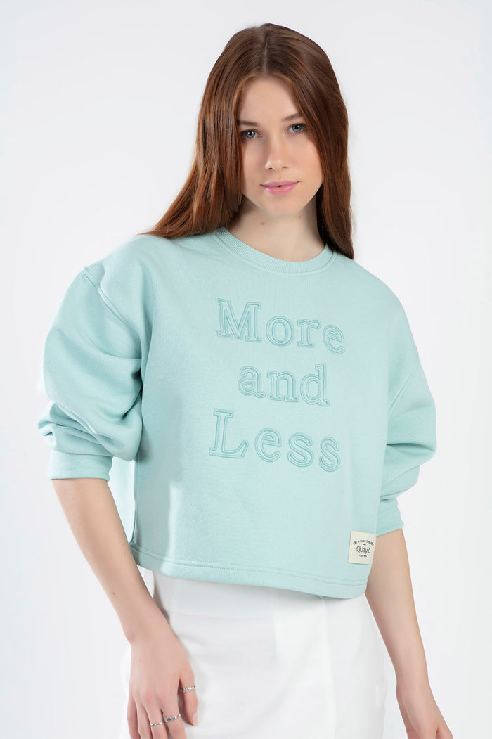 Model in teal More and Less cropped sweatshirt with embossed text and puff sleeves against a white background.