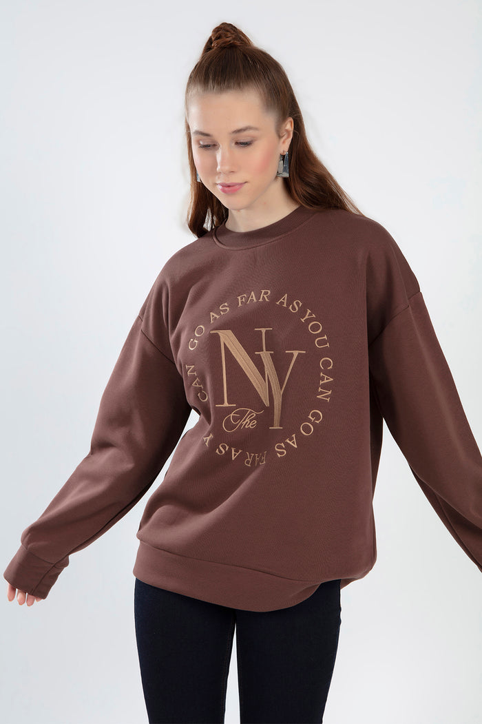 Woman wearing a brown NY graphic sweatshirt with 'Go As Far As You Can' text, featuring an oversized streetwear fit.