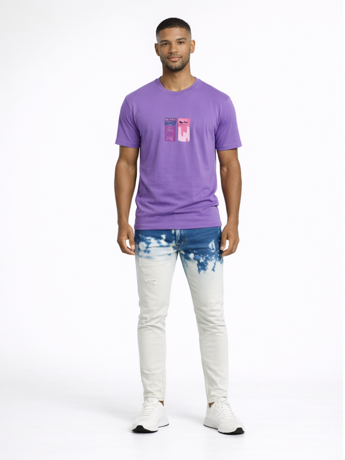 A man stands wearing a purple t-shirt and light blue jeans with a tie-dye pattern, paired with white sneakers.