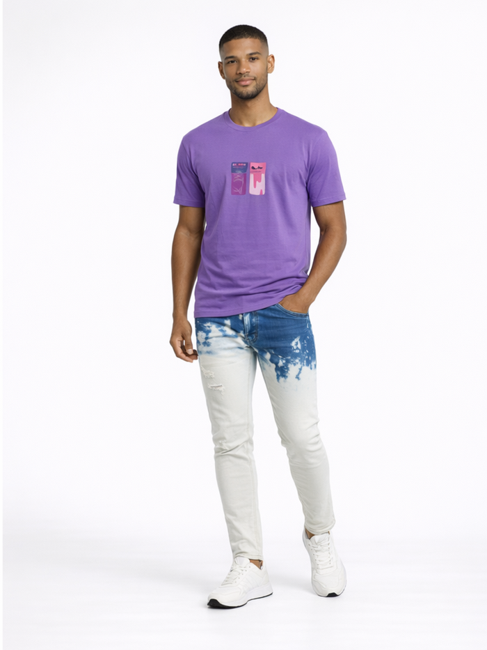 A man stands confidently in a purple t-shirt and light blue, distressed jeans with a tie-dye effect.