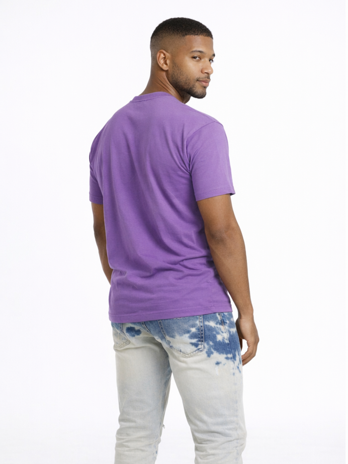 A man with short hair wearing a purple t-shirt and light blue tie-dye jeans, looking over his shoulder.