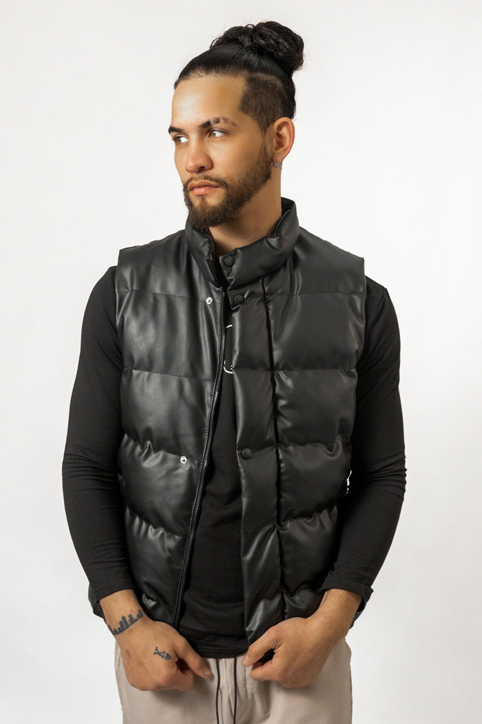 Men's black puffer vest with a high collar and leather-like finish, layered over a long-sleeved streetwear shirt.