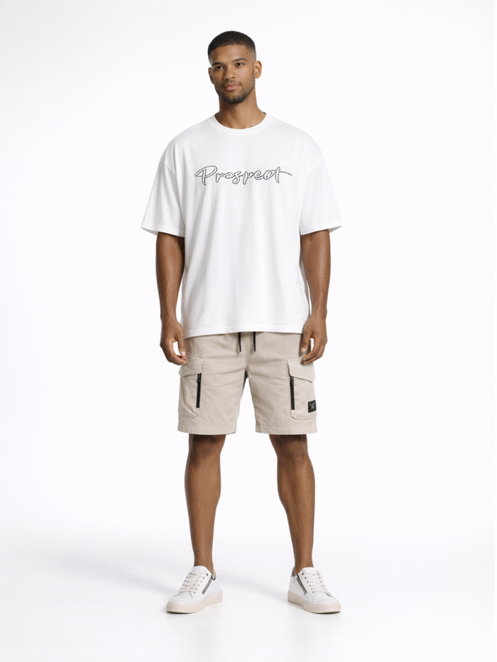 A man stands confidently in a white oversized t-shirt and beige cargo shorts, paired with white sneakers.