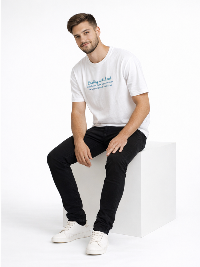 A man sits on a white cube, wearing a casual white t-shirt and black jeans, paired with white sneakers.