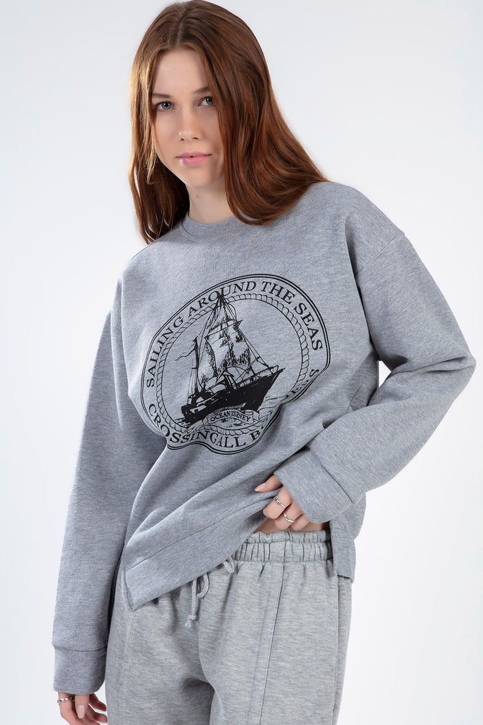 Woman in an oversized light grey streetwear sailing top with a black nautical ship graphic and ribbed cuffs.