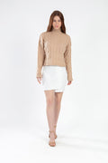 Model wearing Serena cable knit turtleneck sweater in tan with a white mini skirt and heels against a white background.