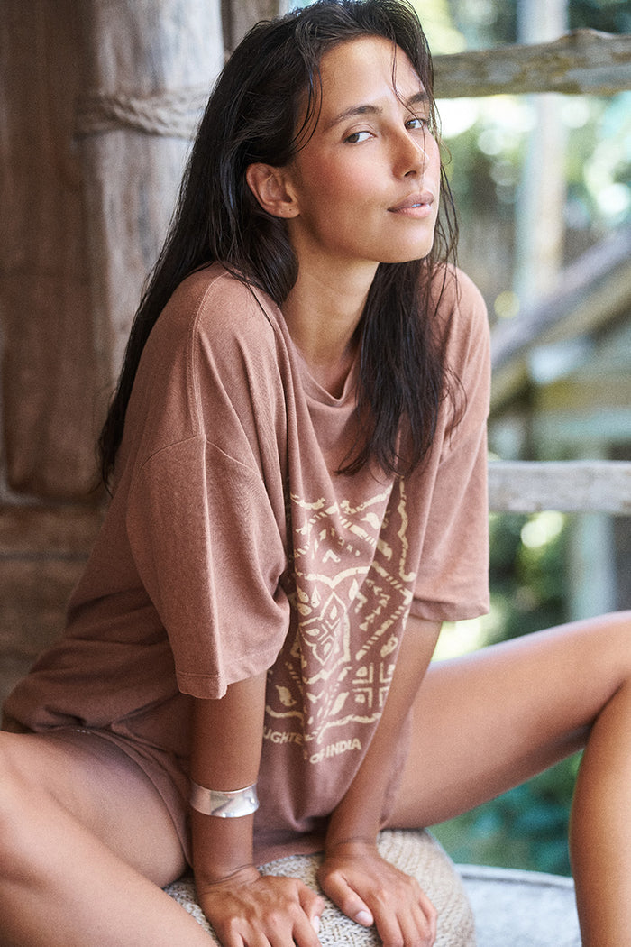 Woman seated outdoors wearing the Daughters of India Shanti Tee in Hazel, an oversized brown block print t-shirt, with silver bangle bracelet
