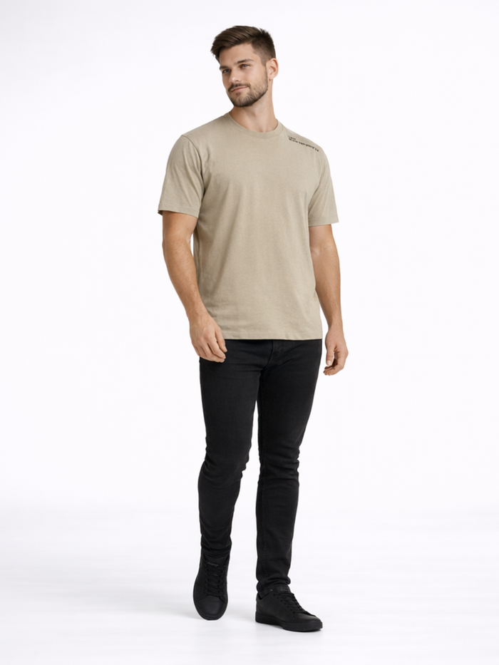 A man wearing a light beige t-shirt and black jeans stands confidently against a plain background.