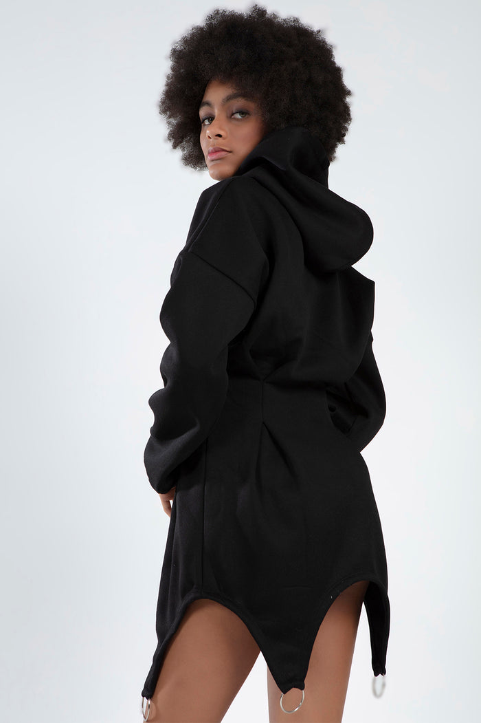 A model with curly hair poses in a black hooded garment featuring unique side cutouts and ring details.