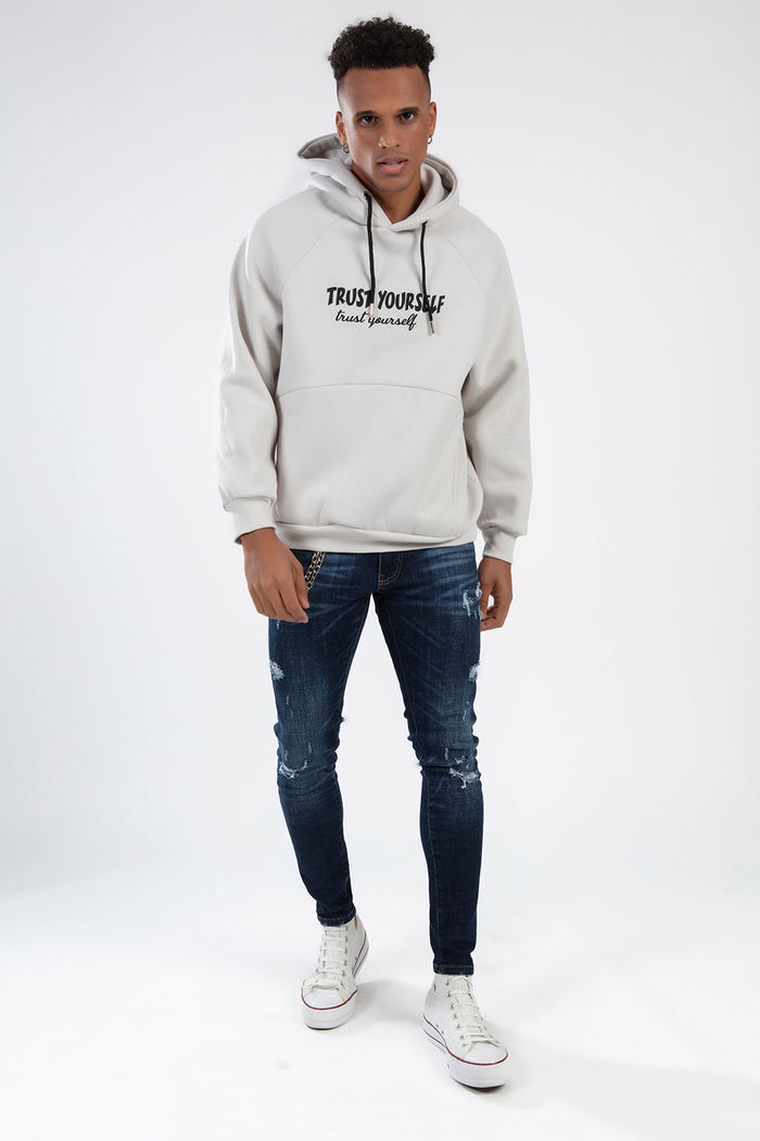 Man wearing Oversized Light Grey Trust Yourself Graphic Hoodie with distressed dark jeans in a streetwear studio shot.