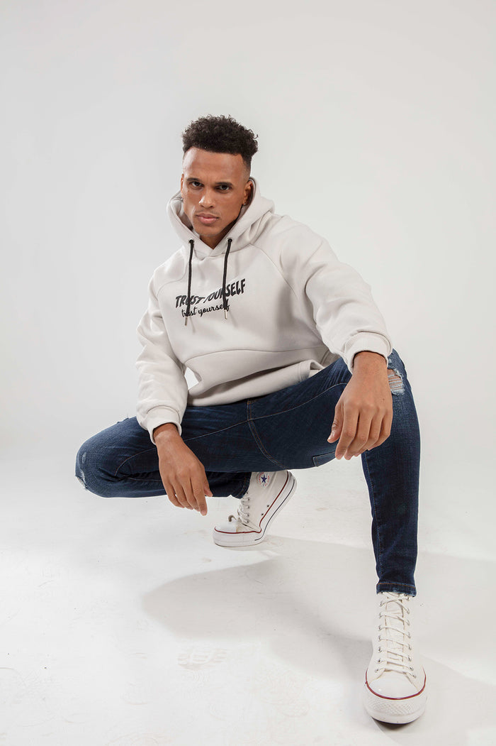 Man in an oversized light grey streetwear hoodie with a 'Trust Yourself' graphic, styled with distressed blue jeans.