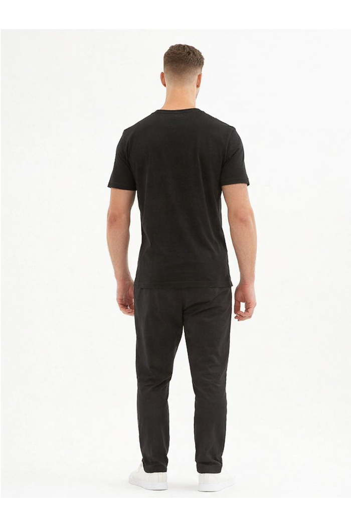 A man wearing a black t-shirt and dark pants stands with his back to the camera, showcasing a casual outfit.
